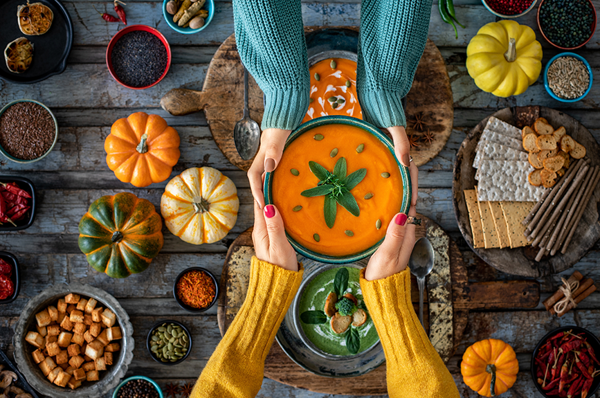 Overhead view of colorful vegetables, pumpkins, and other plant-based ingredients arranged on a wooden table with hands holding a veggie soup bowl, representing vegan cooking and plant-based diet.
