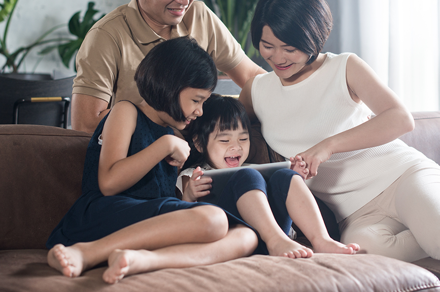 Filipino family with young Gen Alpha children sitting together on a modern couch, parents engaging with kids as they use a tablet device, representing family-oriented digital native generation in contemporary Pinoy home setting.